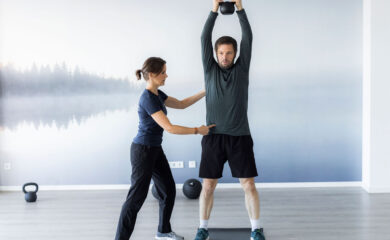 Ein Personal Trainer unterstützt einen Kunden bei einer Kettlebell-Übung mit gestreckten Armen über dem Kopf. Der Trainer korrigiert die Haltung des Kunden für eine optimale Ausführung, während der Kunde konzentriert und stabil steht. Die moderne, helle Trainingsumgebung mit Naturmotiv schafft eine angenehme und motivierende Atmosphäre für das Workout.