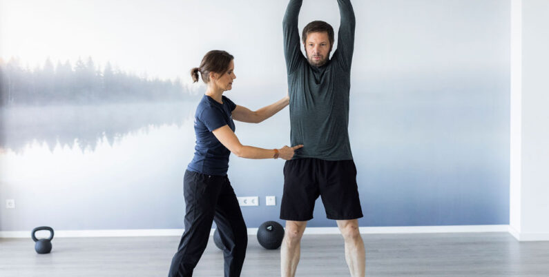 Ein Personal Trainer unterstützt einen Kunden bei einer Kettlebell-Übung mit gestreckten Armen über dem Kopf. Der Trainer korrigiert die Haltung des Kunden für eine optimale Ausführung, während der Kunde konzentriert und stabil steht. Die moderne, helle Trainingsumgebung mit Naturmotiv schafft eine angenehme und motivierende Atmosphäre für das Workout.